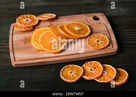 Haufen von Scheiben von frischen und trockenen Orangen auf rechteckigen Holz Schneidebrett auf dunklem Tisch, die als Hintergrund verwendet werden kann Stockfoto