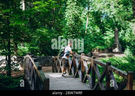 Sofia Park, Uman. Schöner Mann mit Rucksack auf einer Holzbrücke. Männlicher Wanderer auf einer Brücke zwischen grünen Sommerpark. Spaziergang im Sommer grün f Stockfoto