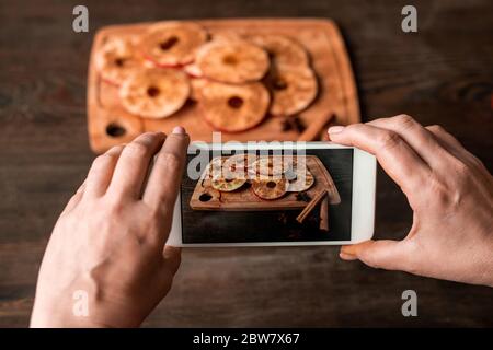 Hände der Hausfrau mit Smartphone Foto von Scheiben von frischem Apfel bestreut mit gemahlenem Zimt auf Holzbrett und Tisch Stockfoto