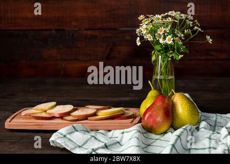 Stillleben Komposition mit Vase aus Wildblumen, Leinen Küchentuch, reife Gartenbirnen und Schneidebrett mit Scheiben von frischem Obst Stockfoto