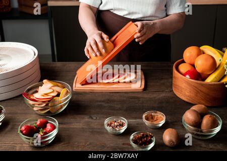 Reife Hausfrau schneidet frische Zitrusfrüchte über Holzbrett von Küchentisch zwischen Schüsseln von Gewürzen, Kiwi, Erdbeeren, Orangen und Banane Stockfoto