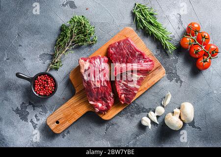 RAW Flap Steak Flanke geschnitten mit Machete, Rock Steak, auf Holz Schneidebrett, mit Kräutern Tomaten Pfefferkörner über grauen Stein Oberfläche Hintergrund vi Stockfoto