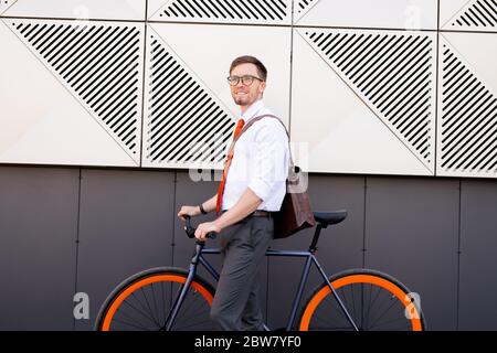 Fröhlicher junger eleganter Mann, der an der Wand moderner Architektur steht, während er Fahrrad benutzt, um nach Hause zu gehen oder nach dem Arbeitstag zu Café Stockfoto