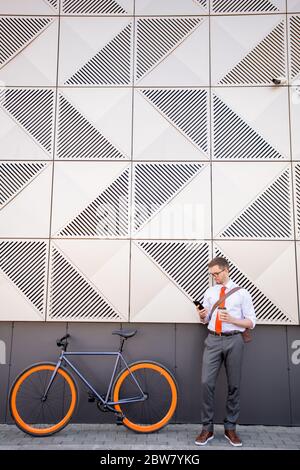 Junger Mann in smarter Freizeitkleidung, der an der Wand eines großen modernen Business-Centers steht und Smartphone benutzt, während er mit dem Fahrrad nach Hause fährt Stockfoto