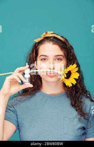 Hübsches junges Weibchen mit Sonnenblume zwischen Nase und Lippen und antiallergischem Nasenspray in der Hand, das vor der Kamera steht Stockfoto