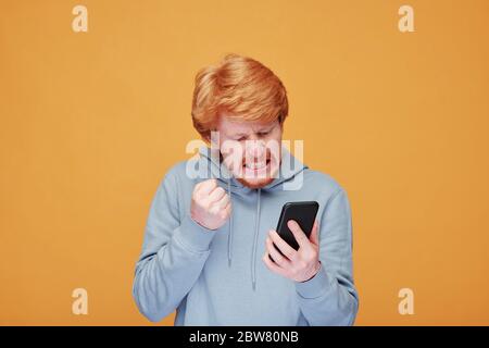 Extrem wütend jungen bärtigen Mann in Casualwear Blick auf Bildschirm des Smartphones, während mit Streit mit jemandem im Video-Chat Stockfoto