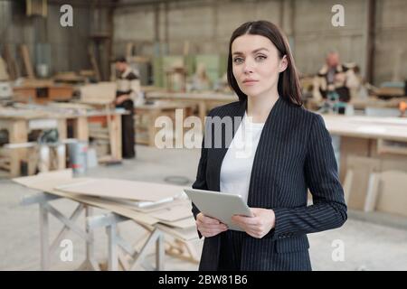 Junge ernsthafte Verkaufsleiter der Möbelfabrik mit Touchpad vor der Kamera, während sie gegen Werkstatt-Innenraum und männliche Arbeiter Stockfoto