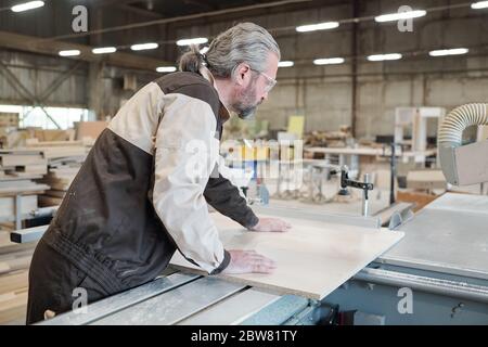 Senior Mann in Arbeitskleidung stehend von großen industriellen Bearbeitungsmaschine und Biegen über Holzbrett, während sie zu schneiden Stockfoto