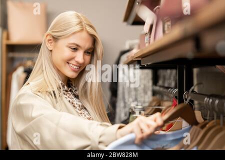 Lächelnde junge blonde Frau mit langen Haaren, die durch neue Mode-Kollektion auf dem Rack schaut, während sie neues Sweatshirt oder Pullover wählt Stockfoto