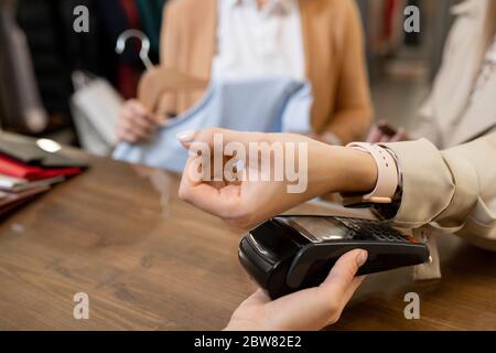 Hand der Kundin mit Smartwatch, die für neue Kleidung in der Boutique oder Bekleidungsabteilung bezahlt, während sie am Schalter steht Stockfoto