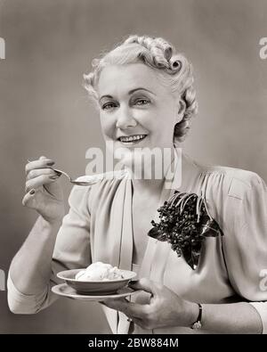 1940ER JAHRE FRAU MITTLEREN ALTERS ESSEN EIN GERICHT EIS TRAGEN KLEID MIT CORSAGE VON KÜNSTLICHEN BLUMEN LÄCHELND SUCHEN TAT KAMERA - F9727 HAR001 HARS LIFESTYLE ZUFRIEDENHEIT ÄLTERE FRAUEN STUDIO-SCHUSS GESUNDHEIT ZU HAUSE LEBEN KOPIE RAUM HALBE LÄNGE DAMEN PERSONEN VERTRAUEN MITTLEREN ALTERS B & W AUGENKONTAKT GLÜCK MITTLEREN ALTERS FRAU FRÖHLICH OLDSTER STILE ERNÄHRUNG LÄCHELN ÄLTESTEN KONSUMIEREN KONSUMIEREN CORSAGE FREUDIGE NAHRUNG STILVOLLE MODE KÜNSTLICHE SCHWARZ-WEISS KAUKASISCHEN ETHNIZITÄT HAR001 ALTMODISCHE TAT Stockfoto