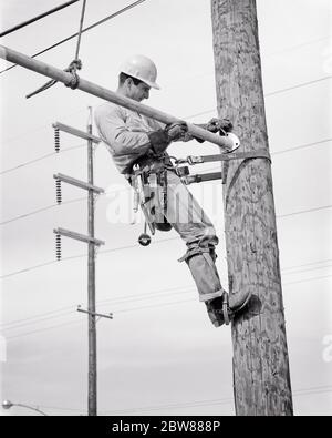 1960S ELEKTRISCHE LINEMAN AUF VERSORGUNGSMAST MIT SICHERHEITSGURT UND GAFFEN ODER SPOREN AN SEINEN FÜSSEN MIT EINEM HARTEN HUT UND WERKZEUGGÜRTEL GESICHERT - I3838 HAR001 HARS BLAUEN KRAGEN FÄHIGKEITEN BERUF FÄHIGKEITEN SEINE MANUELLE UND NIEDRIGE WINKEL ARBEIT BESCHÄFTIGUNG BERUFE VERSORGUNGSUNTERNEHMEN MITARBEITER LINEMAN ODER HANDEL BLAUE JEANS MITTE ERWACHSENE MITTE ERWACHSENE MANN PRÄZISION GESICHERT SPOREN SCHWARZ UND WEISS KAUKASISCHEN ETHNIZITÄT GITTER HAR001 INSTALLATION ARBEIT ALTMODISCHEN ÖFFENTLICHEN VERSORGUNGSUNTERNEHMEN Stockfoto