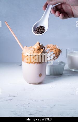 Man's Hand hinzufügen Schokolade Chips zu einem Dalgona Kaffee auf weißem Hintergrund. Koreanischer Trenddrink. Vertikales Bild. Stockfoto