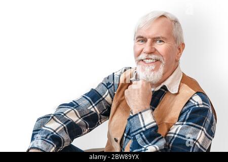 Freistil. Senior bärtig stilvollen Mann sitzt isoliert auf weiß berühren Kinn lächeln freudig Nahaufnahme Stockfoto