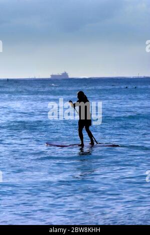 Der ältere Mann ist silhouetted, als er seine Bretter über Waikiki Bay paddelt. Schiff verlässt im Hintergrund den Hafen. Abend Licht Silhouetten Paddler. Stockfoto