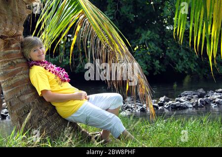 Attraktive Weibchen entspannt sich an einer schiefen Palme auf der Big Island von Hawaii. Sie trägt eine orchideenkette, während Palmwedel tropischen Seerosken umrahmen Stockfoto