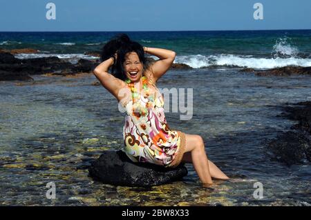 Hawaiianerin sitzt auf einem Felsen im flachen Wasser und feiert, indem sie ihre Haare hochwirft und posiert. Anaehomalu Bay an der Kohala Küste der Big Isla Stockfoto