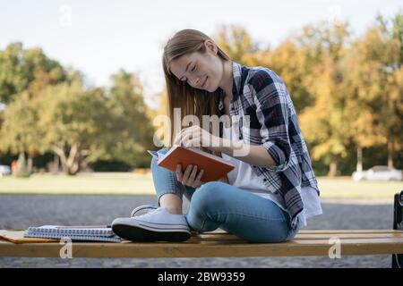 Schöne Frau, die ein Buch liest, das auf Bank sitzt. Student Studium, Sprache lernen, Prüfungsvorbereitung, Arbeitsprojekt im Park. Bildungskonzept Stockfoto