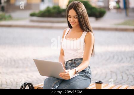 Attraktive junge Frau Laptop draußen auf der Bank im Sommer. Frau, die im Freien online arbeitet. Allein. Schöne Brünette weiblich. Studierendenstudium Stockfoto