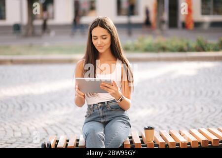 Schöne Mädchen verwenden Tablet im Freien. Frau allein auf der Bank sitzend. Frau, die online arbeitet Stockfoto