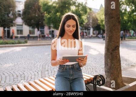 Schöne Mädchen verwenden Tablet im Freien. Frau allein auf der Bank sitzend. Frau, die online arbeitet Stockfoto