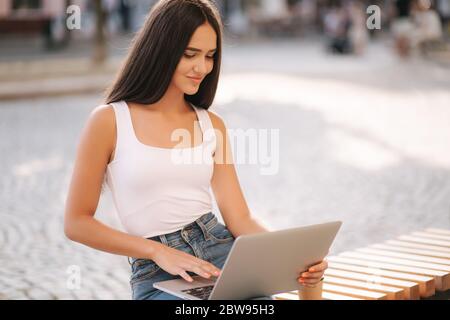 Attraktive junge Frau Laptop draußen auf der Bank im Sommer. Frau, die im Freien online arbeitet. Allein. Schöne Brünette weiblich. Studierendenstudium Stockfoto