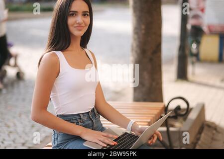 Attraktive junge Frau Laptop draußen auf der Bank im Sommer. Frau, die im Freien online arbeitet. Allein. Schöne Brünette weiblich. Studierendenstudium Stockfoto