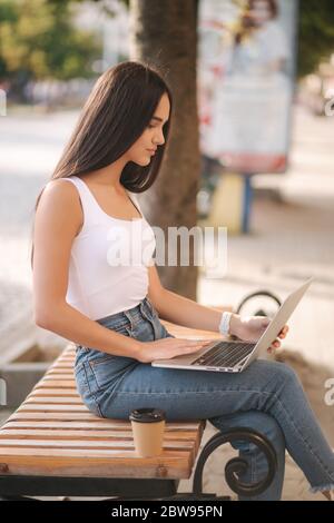 Attraktive junge Frau Laptop draußen auf der Bank im Sommer. Frau, die im Freien online arbeitet. Allein. Schöne Brünette weiblich. Studierendenstudium Stockfoto