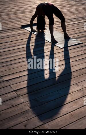 Fit Mädchen demonstriert Yoga Asana Urdha Dhanurasana auf dem Holzboden im Freien im Licht und langen Schatten des Sommers Sonnenuntergang Stockfoto