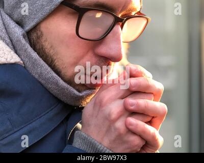 Nahaufnahme Porträt des Mannes, der ihre Hände mit Atem in sonnigen Wintertag wärmt, Brillen beschlagen, Handschuhe zu Hause vergessen. Mann Hände bei kaltem Wetter. Stockfoto