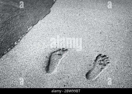 Ein sanfter Wasserrausch bedroht eine einzige Reihe von Fußabdrücken an einem Strand auf der Insel Oahu. Das Bild ist schwarz-weiß Stockfoto