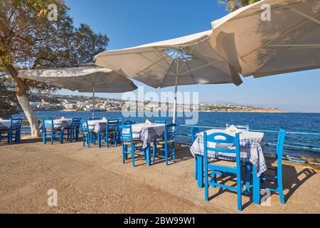 Ein leeres kretisches Restaurant mit blauen Stühlen und Tischen mit Sonnenschirmen am Meer, im Hintergrund die Stadt Rethymno Stockfoto