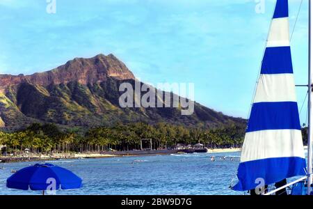 Diamond Head wird von einer blauen Umberella und einem blau-weißen Segelboot eingerahmt. Schwimmer genießen das hawaiianische Wasser vor Waikiki Beach Stockfoto