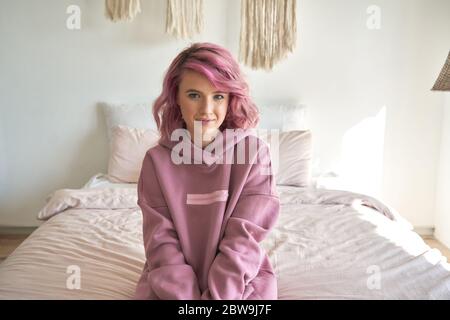 Lächelnd Hipster gen z jugendlich Mädchen mit rosa Haar tragen Kapuzenpullover sitzen auf dem Bett, Porträt. Stockfoto