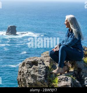 USA, Kalifornien, Big Sur, Frau, die am Rand der Klippe sitzt und die Aussicht betrachtet Stockfoto