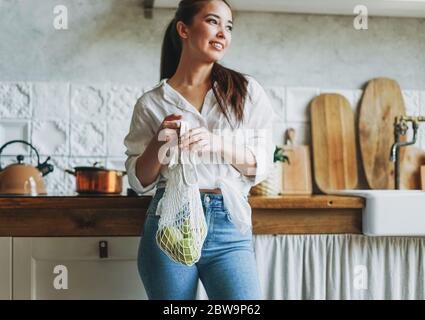 Junge asiatische Frau in weißen Hemd halten gestrickte Stofftasche Shop mit grünen Äpfeln in Händen auf Küche Stockfoto