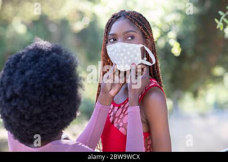 Schöne afroamerikanische Mädchen hilft ihrem Freund, eine Gesichtsmaske auf setzen. Gesichtsschutz ist wichtig, um vor Viren, einschließlich COVID-19, zu schützen Stockfoto