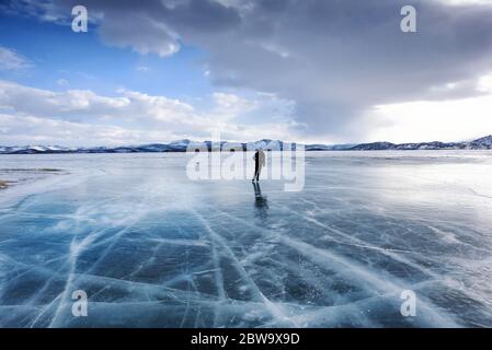 Baikalsee, Russland - 13. März 2020: Mann Eislaufen auf dem gefrorenen Baikalsee mit schneebedeckten Bergen im Hintergrund Stockfoto