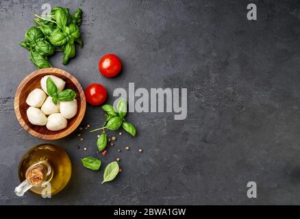 Zutaten für Caprese Salat. Rote Tomaten, Olivenöl, frische Kräuter-Basilikumblätter und Mozzarella-Kugeln über schwarzem Steintisch. Stockfoto