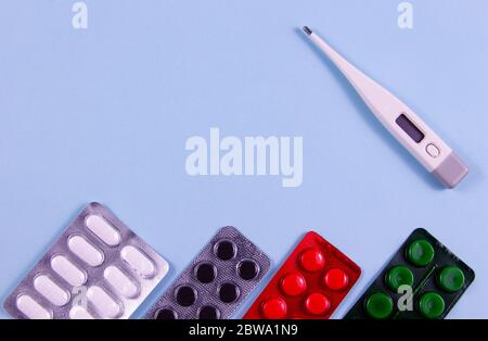 Weiße grüne schwarze und rote Pillen in Blisterpackungen und elektronisches Thermometer auf blauem Hintergrund. Foto von oben. Objekte befinden sich auf sid Stockfoto