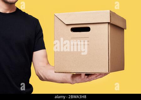 Liefermann in schwarzer Uniform halten Pappschachtel auf gelbem Hintergrund. Konzept des Delivery Service Stockfoto
