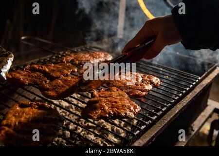 Abends ein Barbecue-Grill, auf dem leckere saftige Steaks über offenem Feuer gegrillt werden, eine Hand hält die Zange und dreht das Fleisch um. Nahaufnahme, weich Stockfoto