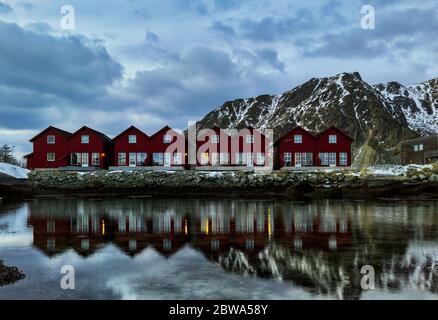 Eine horizontale Aufnahme einer symmetrischen Reihe von klassischen roten Rorbu neben schneebedeckten Bergketten, die Wasser in Ballstad, Lofoten Island, Norwegen reflektieren Stockfoto