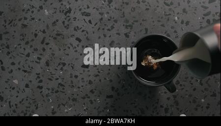 Gießen Sie Sahne in schwarzen Kaffee in schwarzer Tasse auf Beton Draufsicht im Hintergrund Stockfoto