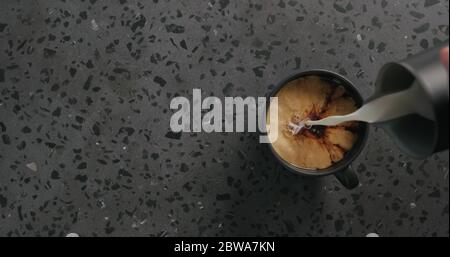 Gießen Sie Sahne in schwarzen Kaffee in schwarzer Tasse auf Beton Draufsicht im Hintergrund Stockfoto
