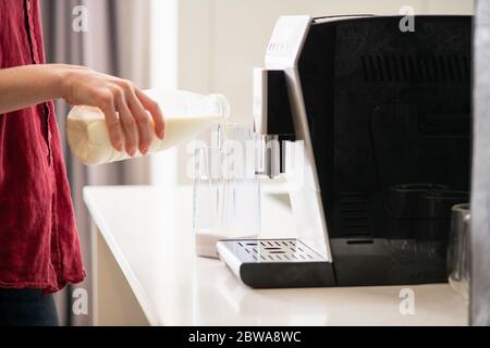 Weibliche Hände gießen frische Milch aus Kunststoffflasche Kaffeemaschine transparenten Behälter für Heißgetränk, Cappuccino, Latte, Macchiato zu Hause vorzubereiten Stockfoto