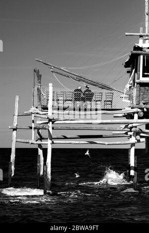 Ein Paar von hinten gesehen, auf der Brücke eines Trebuchets in San Vito Chietino, Abruzzen, Italien, über das Meer, während Möwen unter dem Meer fliegen Stockfoto