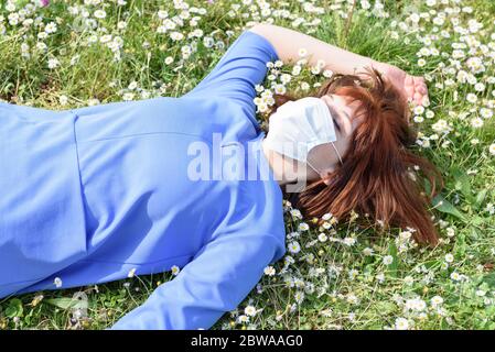 Mädchen auf der blühenden Wiese der Gänseblümchen mit chirurgischer Maske. Ein Mädchen in einem blühenden Garten liegt auf dem Gras Stockfoto