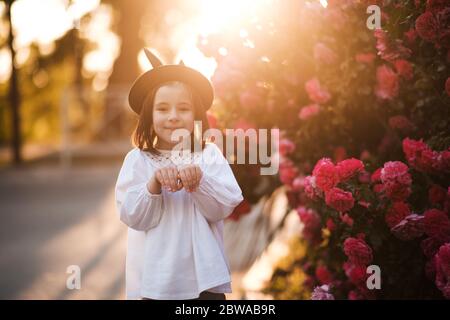 Lächelnd niedliches Baby Mädchen 3-4 Jahre alt das Tragen stilvolle Sommerkleidung und Spaß über Rosen im Hintergrund draußen in der Stadt Straße. Betrachten der Kamera. Stockfoto