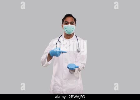 Arzt zeigt auf leere offene Hand vor der Brust Tragen von medizinischen Maske und Handschuhe isoliert. Indian Man Doctor Advertisment Konzept Stockfoto